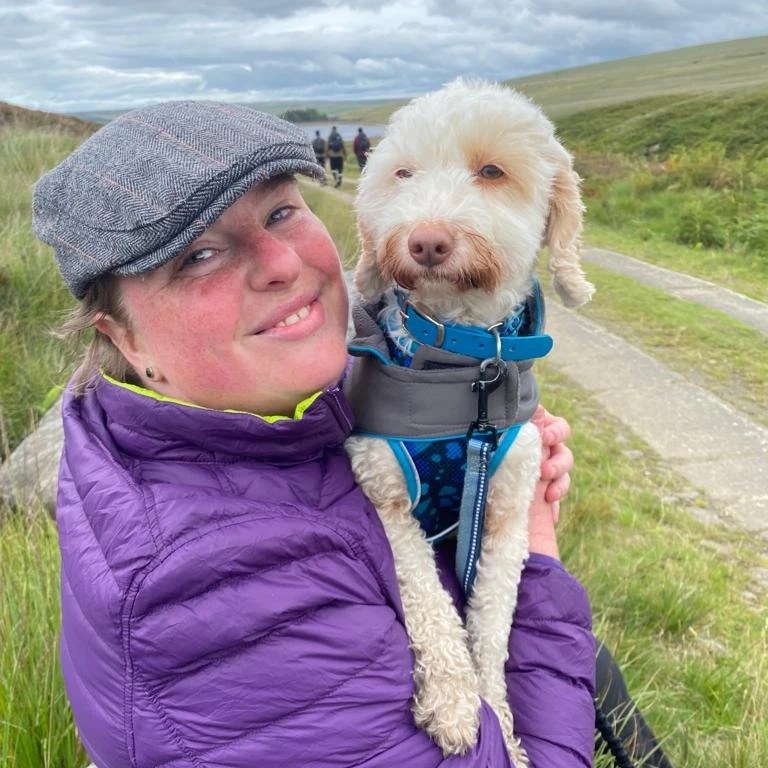 Becci is wearing hiking gear and a flat cap. She is carrying Mossy the white cockapoo, who is wearing her grey coat and lead. Becci is on the moors and you can see a reservoir behind her. She is smiling because she feels motivated.